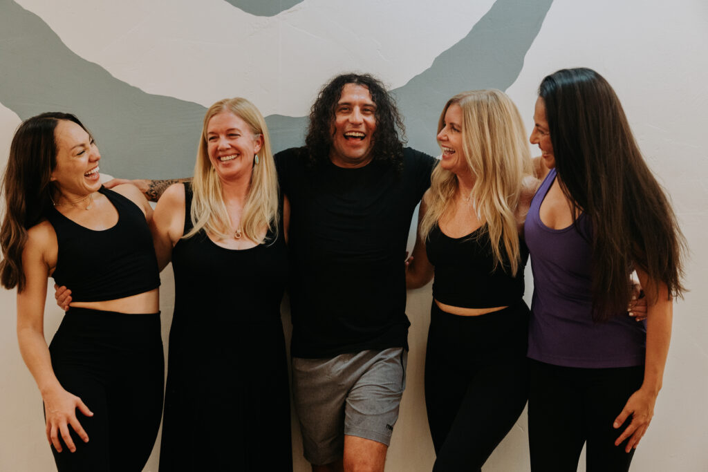 Five instructors laughing near the the company logo on the wall